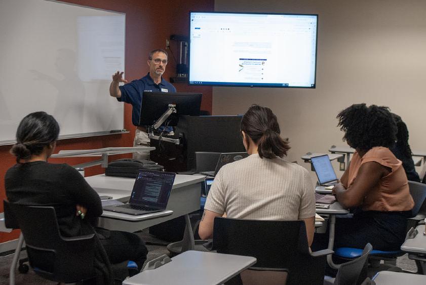 Four people sit in a semicircle in a classroom listening to speaker and watching a screen